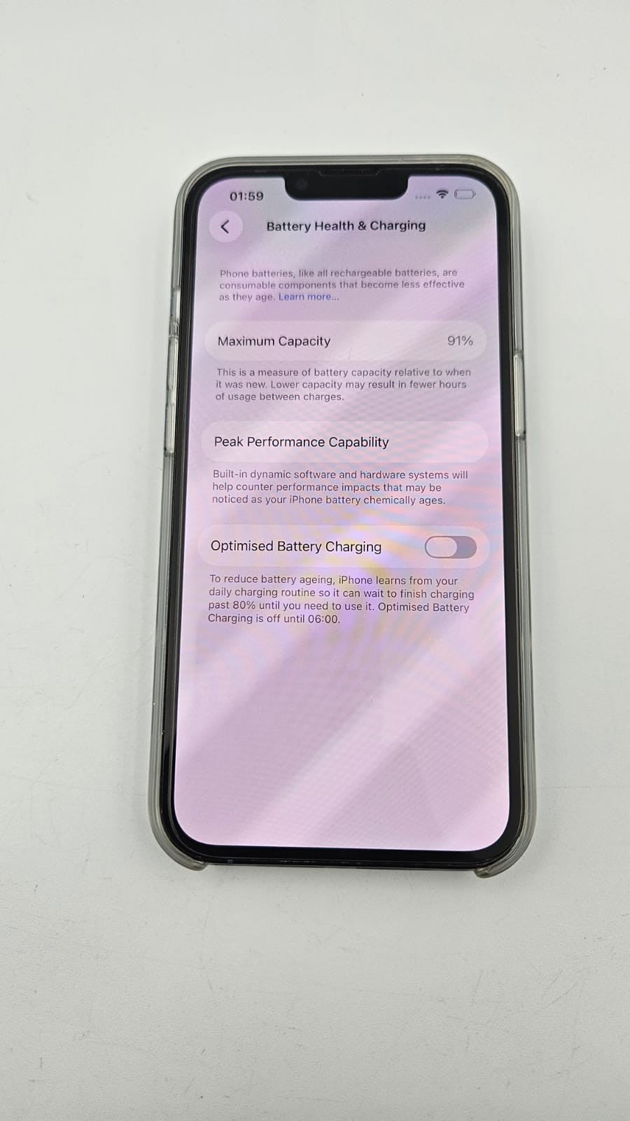 A black iPhone 13 pro lying flat on a light gray surface, displaying the iOS Battery Health & Charging settings screen.