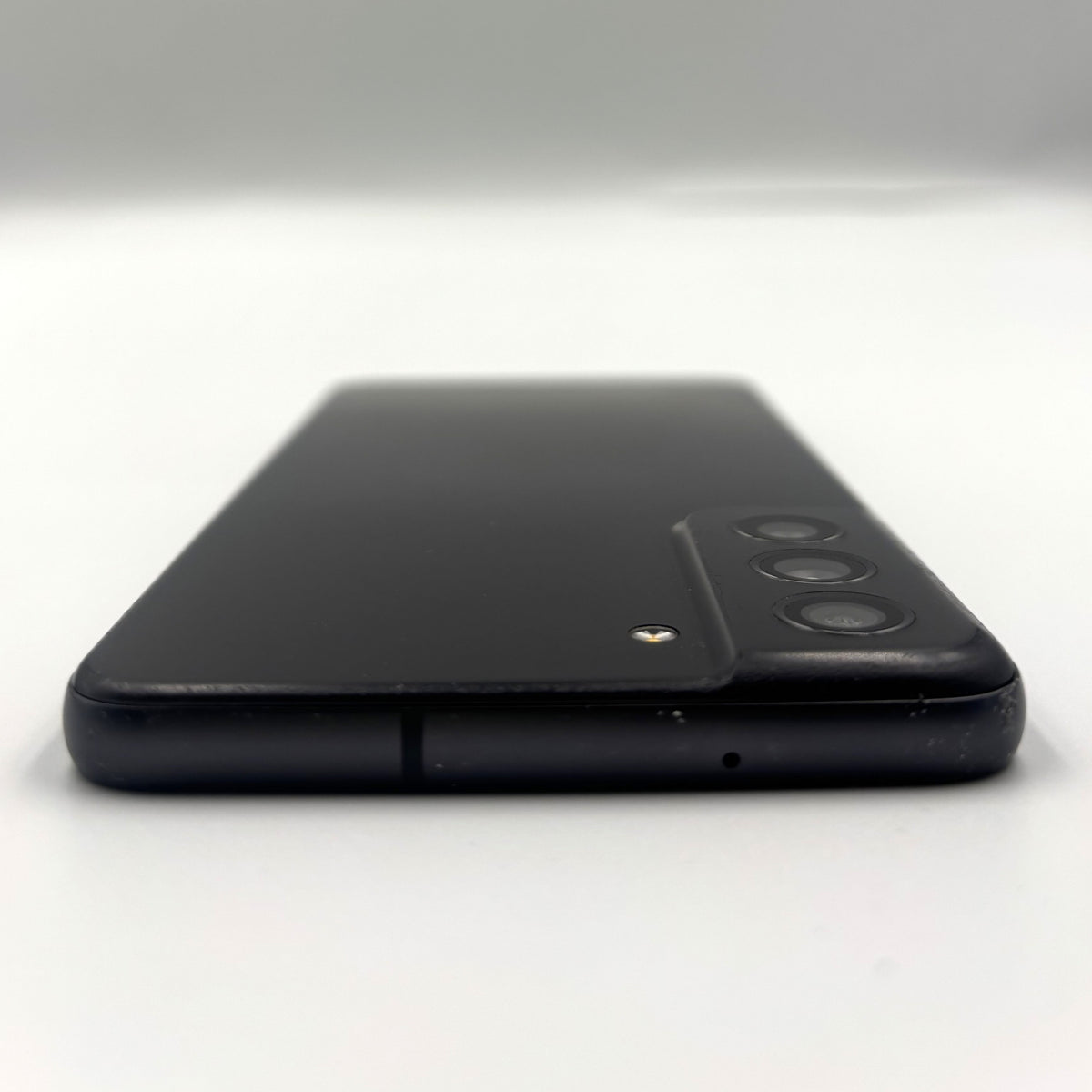 A black smartphone shown from the back, photographed on a plain light gray to white studio background. The back panel is matte black with a raised camera module containing three circular lenses and a small gold accent near the top.