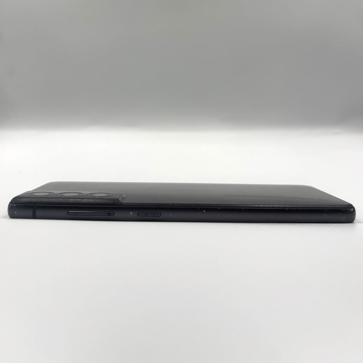 A black Samsung S21 Fe shown from the side, displaying its slim profile, camera cutout, and side buttons against a neutral gray studio background.