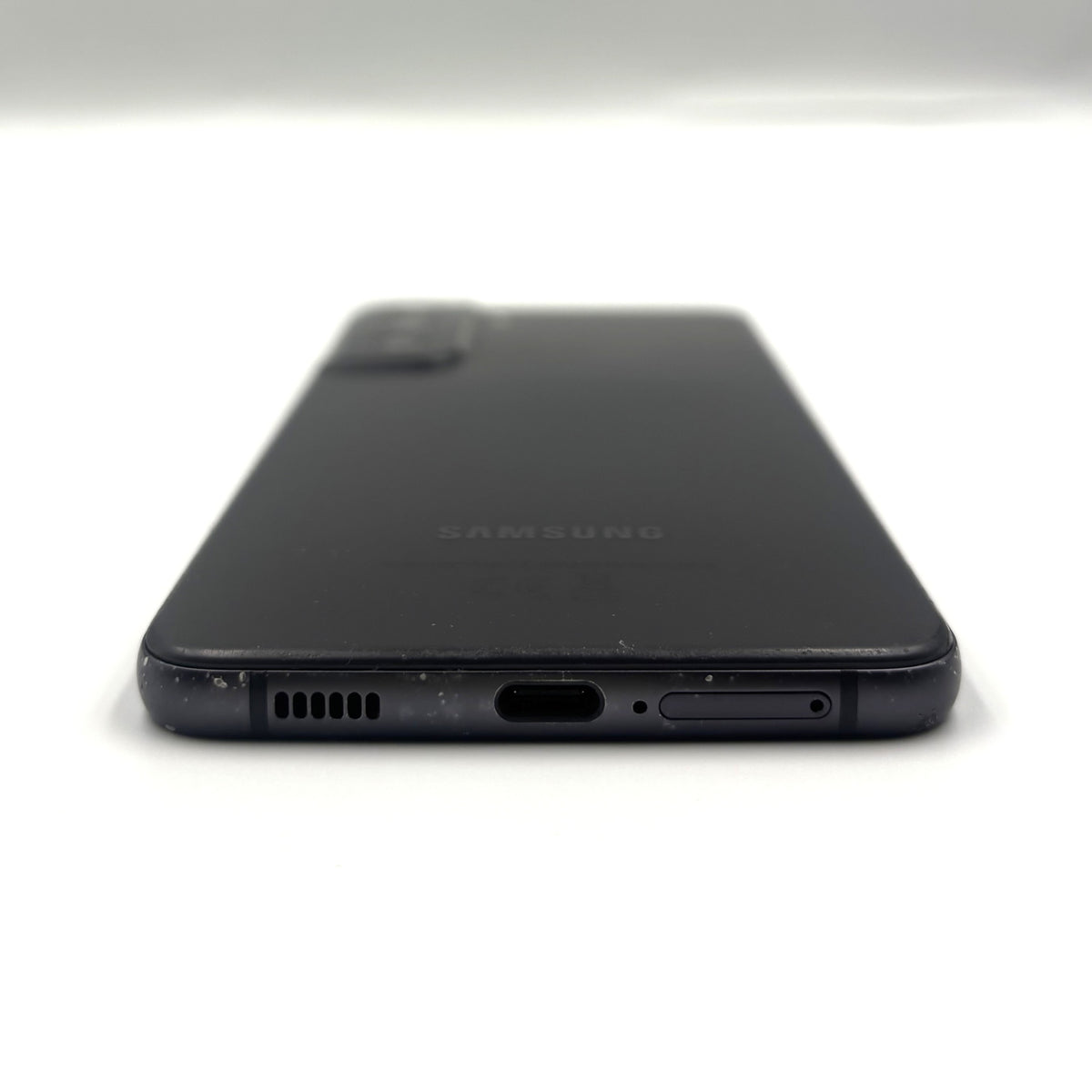 The back of a black Samsung smartphone, showing the camera module, speaker grille, charging port, and side button, photographed on a white background.