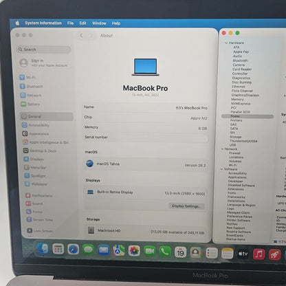 A silver Apple MacBook Pro laptop open and in use, showing the macOS desktop with several application windows open, photographed against a plain light wall.